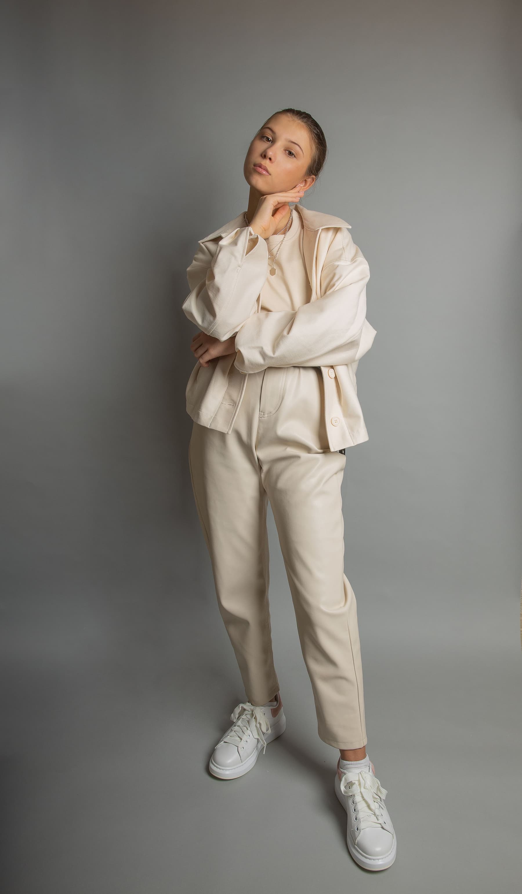 A woman in a cream coat, seated low against a plain grey wall.